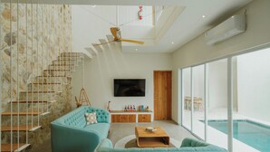 A modern living room with a stone wall, a staircase, a teal sofa, a wooden coffee table, a flat-screen TV, and a large window overlooking a pool.