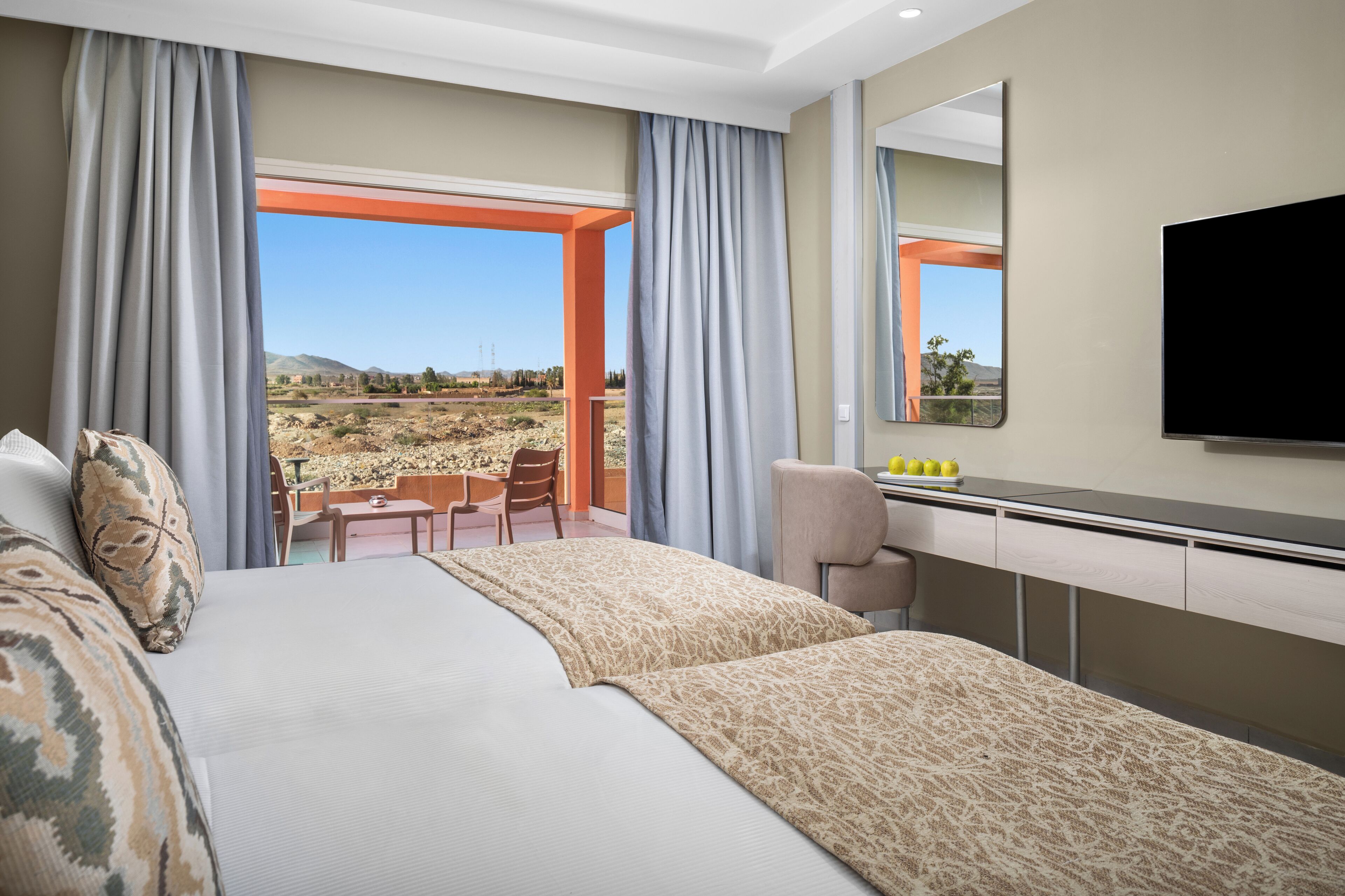 Ein Hotelzimmer mit einem großen Bett, einem Schreibtisch, einem Sessel und einem Fernseher. Ein Balkon bietet Blick auf Wüste und Berge.
