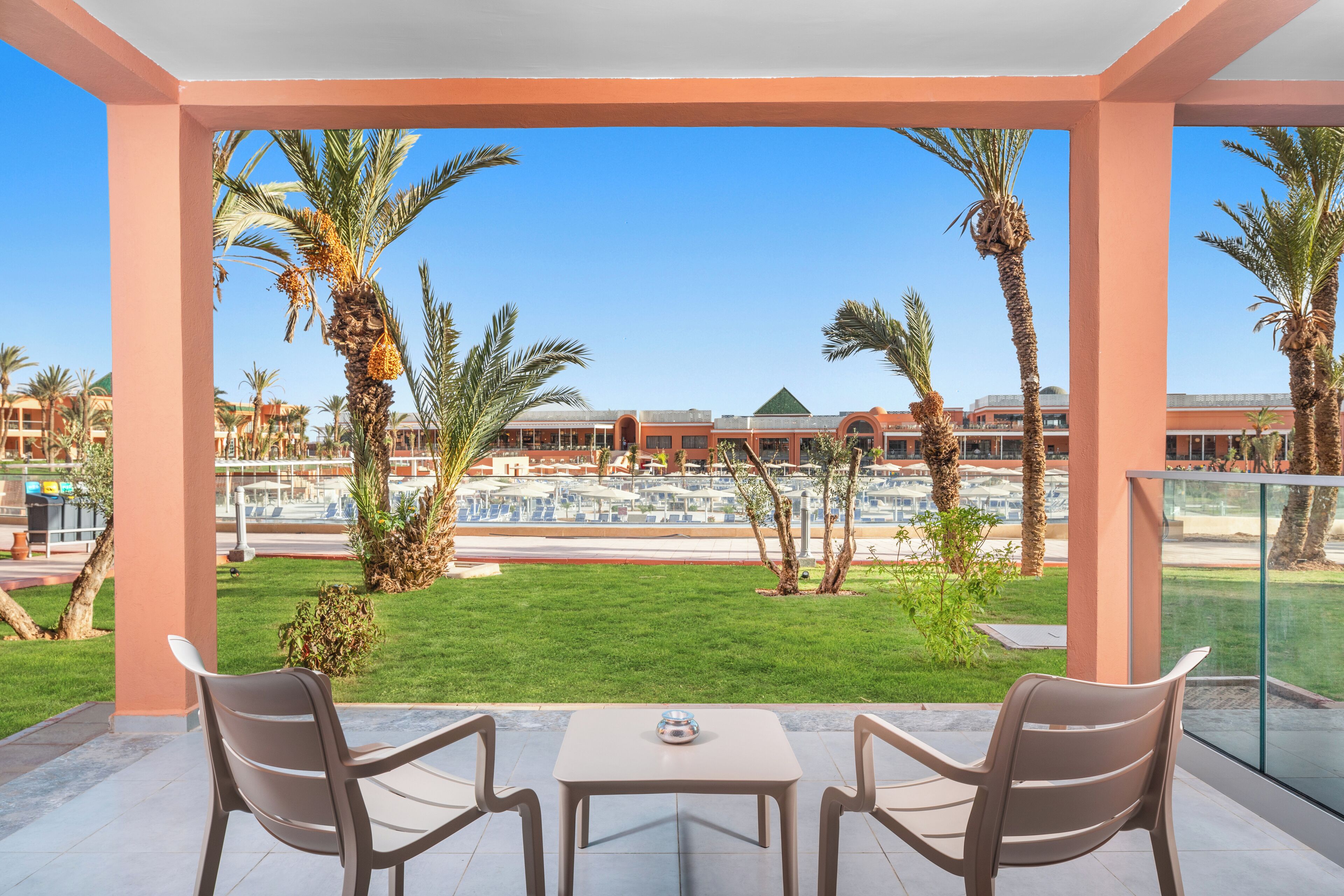 Ein überdachter Außenbereich mit zwei Stühlen und einem Tisch, mit Blick auf einen angelegten Garten mit Palmen und im Hintergrund Hotelgebäuden.