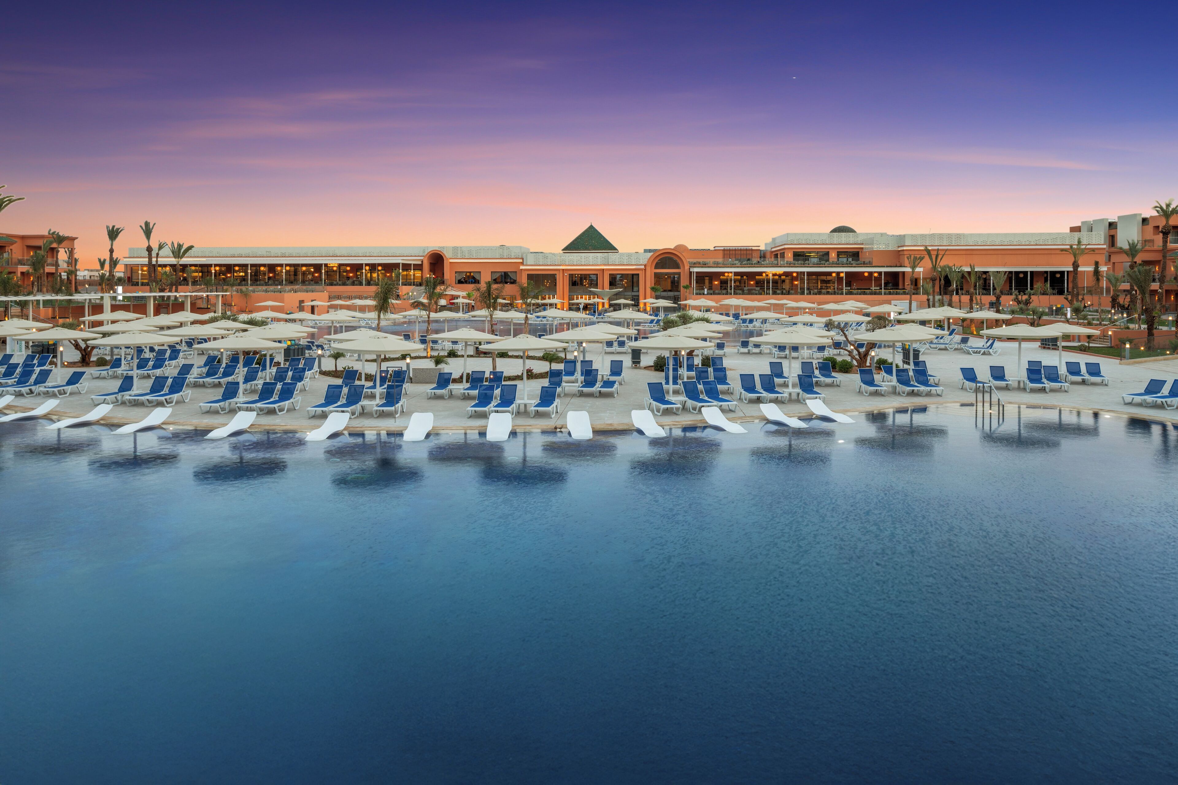 Un vaste espace piscine doté de nombreuses chaises longues et de parasols, entouré d’un bâtiment d’hôtel se distinguant par son toit vert.
