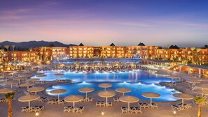 Un resort con una amplia piscina rodeada de tumbonas y sombrillas, varios edificios de hotel y palmeras.