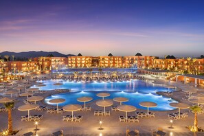 Un resort con una amplia piscina rodeada de tumbonas y sombrillas, varios edificios de hotel y palmeras.