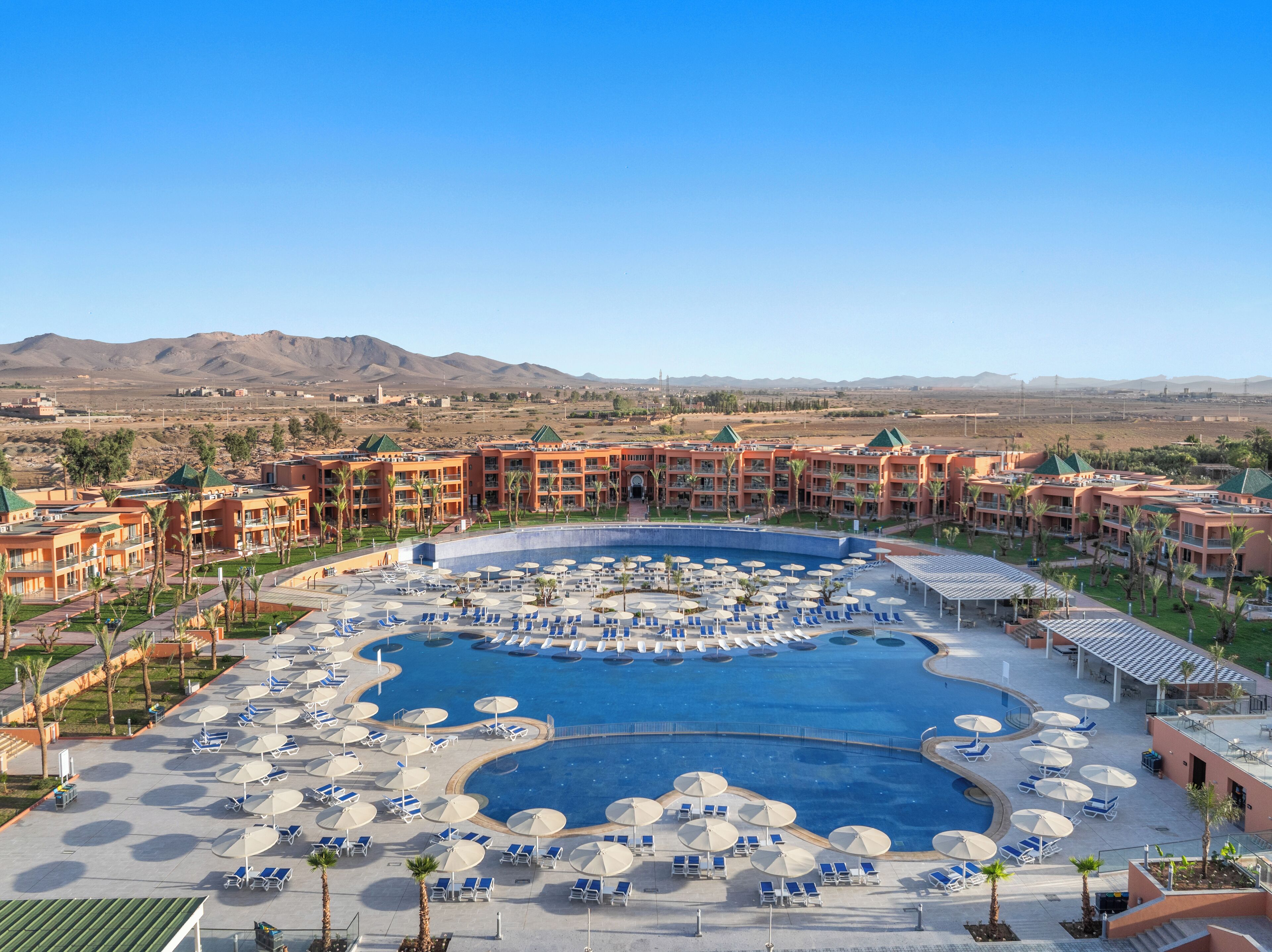 Un complexe hôtelier doté d’une grande piscine centrale entourée de transats et de parasols, de plusieurs bâtiments d’hôtel avec des balcons, et d’un paysage désertique en arrière-plan.