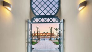 An open doorway leading to a building with a tiled pathway, palm trees, and a view of a hotel with a green roof and multiple floors.