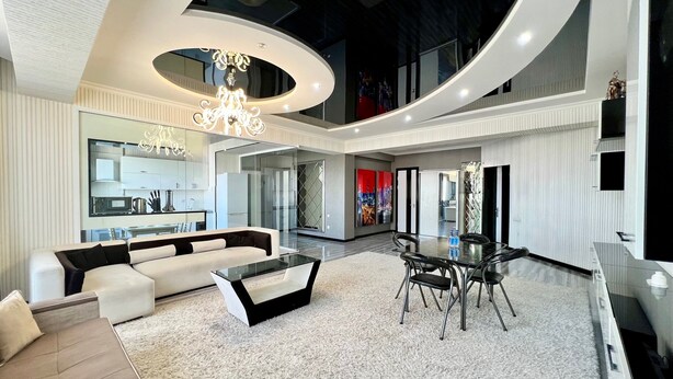 A modern living room with a sofa, coffee table, and dining table. There is a kitchen in the background with a refrigerator and cabinets. - Golden house (Tashkent)