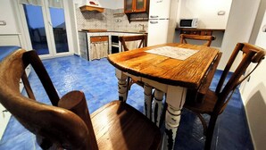 Un coin repas avec une table et des chaises en bois, une cuisine équipée d’un réfrigérateur et d’un four à micro-ondes, et une vue à travers une porte vitrée.