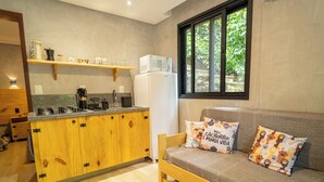 A compact kitchen with wooden cabinets, a built-in stove, and a refrigerator. There is a window providing natural light and a sofa with decorative pillows. - Bom pra Cachorro (Morro de São Paulo)