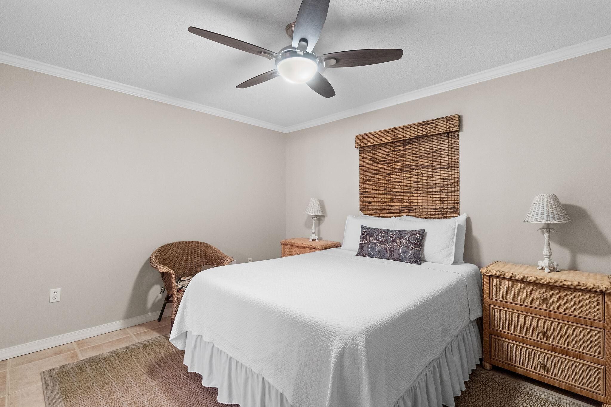 A bedroom with a bed, a ceiling fan, a wicker nightstand, a chair, and a lamp.