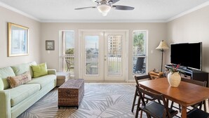 Un salon avec un ventilateur de plafond, un canapé, une table basse, une table à manger avec des chaises, un téléviseur et une fenêtre avec des stores.