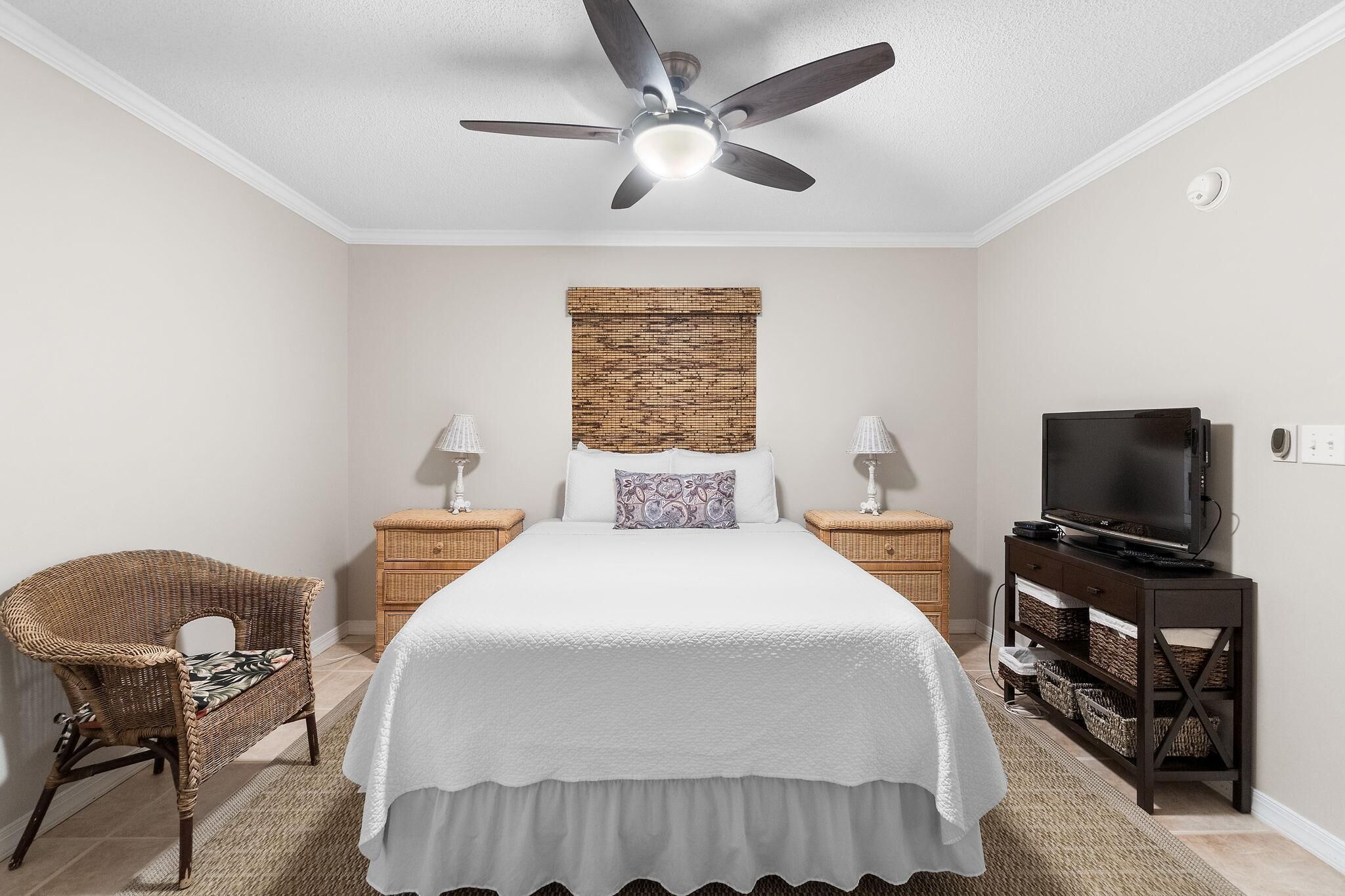 A bedroom with a bed, a ceiling fan, a television, a wicker chair, and a nightstand.