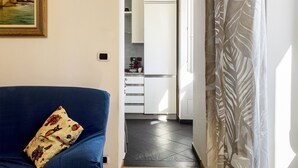 Un salón con un sofá azul y un cojín estampado, que da a una cocina con armarios blancos y suelo de baldosas.