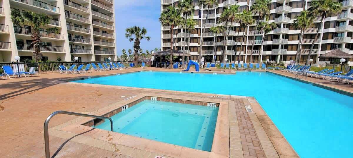 Una piscina con tabla de clavados, rodeada de sillas de descanso y palmeras, con edificios de varios pisos al fondo.
