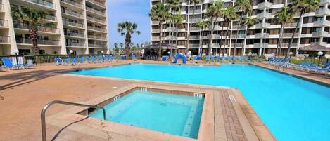 Una piscina con tabla de clavados, rodeada de sillas de descanso y palmeras, con edificios de varios pisos al fondo.