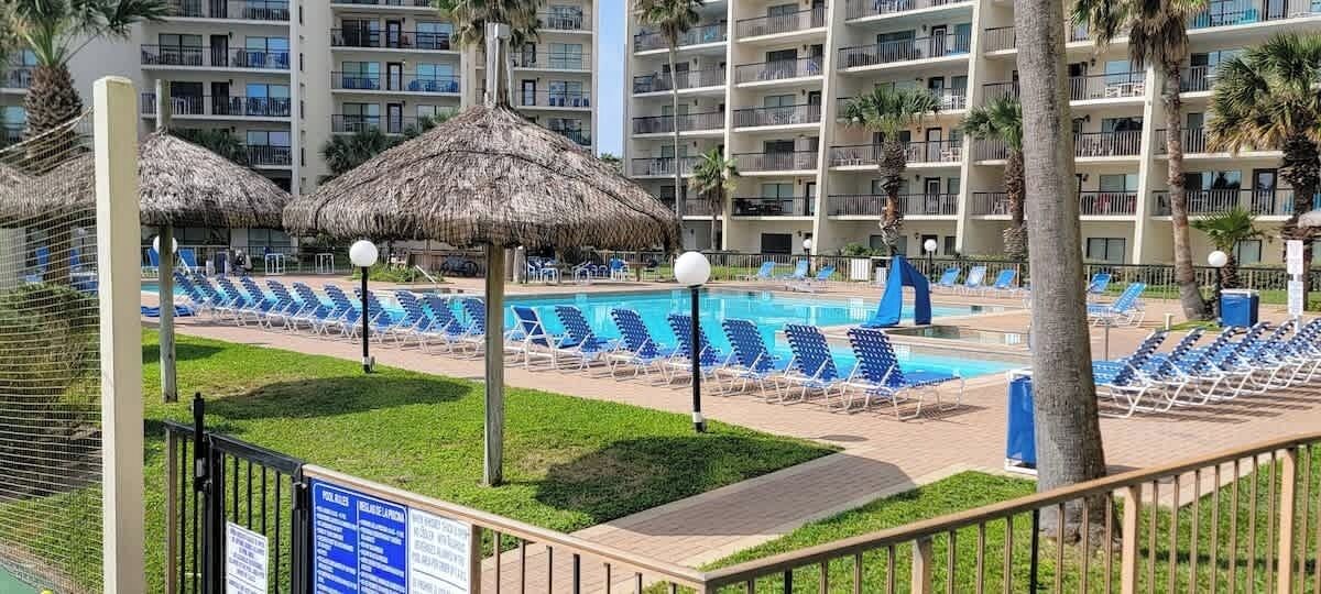 A pool area with blue lounge chairs and thatched umbrellas, surrounded by a fence and palm trees.