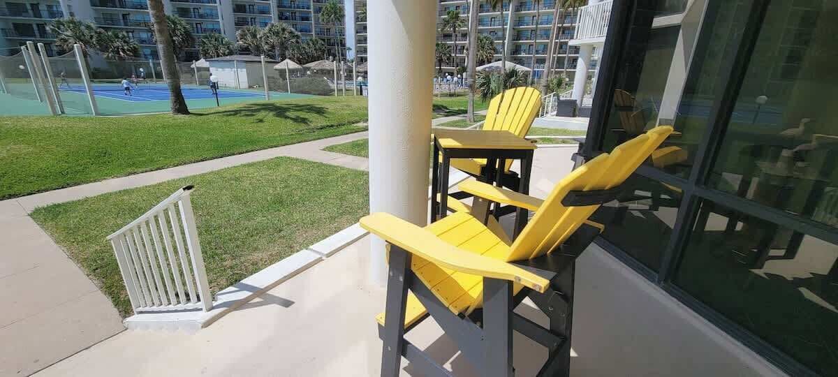 A balcony with yellow chairs and a white railing, overlooking a tennis court and a pool area.