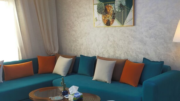 Una sala de estar moderna con un sofá verde azulado, cojines blancos y anaranjados, una mesa de centro de vidrio y un sillón blanco.