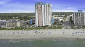 A beachfront hotel with multiple floors, a sandy beach with umbrellas, and a view of the ocean.
