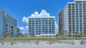 A modern hotel with a central glass facade, surrounded by beach vegetation and other buildings.