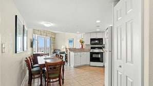 A modern kitchen with white cabinets, a dining area with a wooden table and chairs, and a view of the outdoors through large windows.