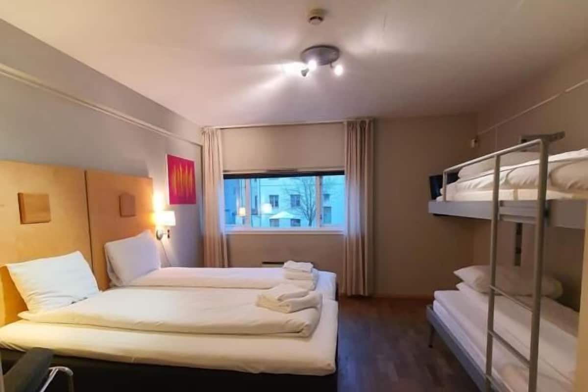 Una habitación de hotel con cama, ventana con cortinas, literas y mesita de noche con lámpara.