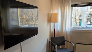 A room with a flat-screen TV mounted on the wall, a chair, a lamp, and a window with curtains.