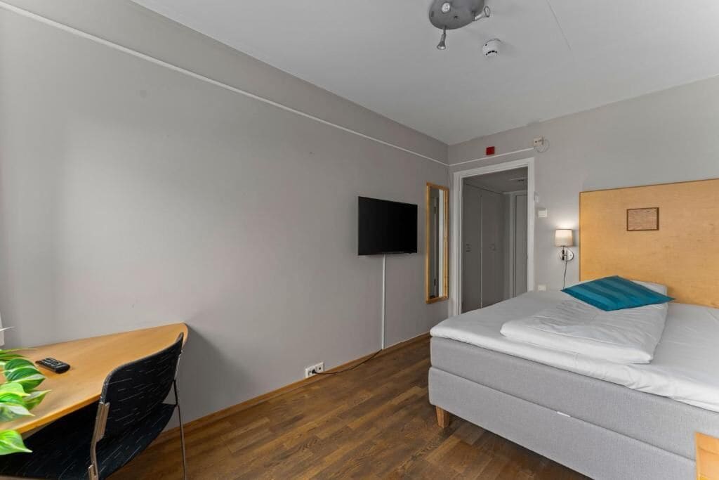 A hotel room with a bed, a desk with a chair, a television mounted on the wall, and a wooden floor.