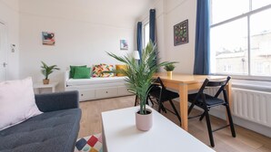 A modern, well-lit room with a sofa, a dining table with chairs, a small kitchen area, and large windows with curtains. - Split-level spacious family apartment (London)