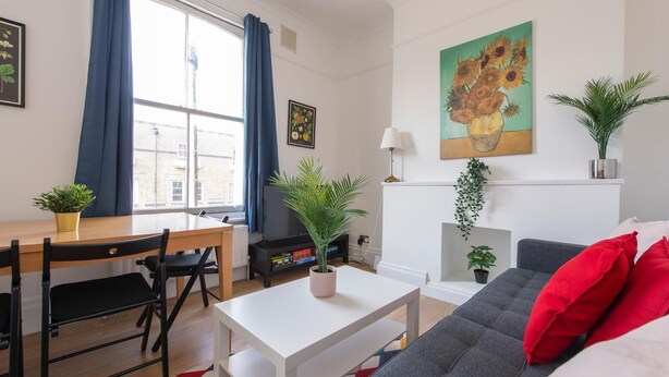 A modern living room with a sofa, a dining table with chairs, a TV, and a fireplace. There are potted plants and artwork on the walls. - Split-level spacious family apartment (London)