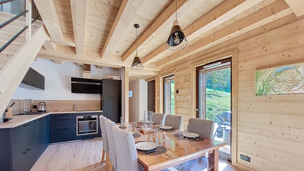 Eine moderne Küche mit Einbauofen und einem schwarzen Kühlschrank. Ein Essbereich mit Holztisch und Stühlen lädt zum Verweilen ein, und ein großes Fenster bietet Blick ins Freie.