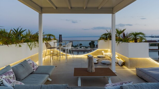 A covered outdoor seating area with a view of the sea, featuring a sofa, chairs, a table, and ambient lighting. - Terrace Jacuzzi - Tropicalia by Wonderful Italy (Aci Castello)