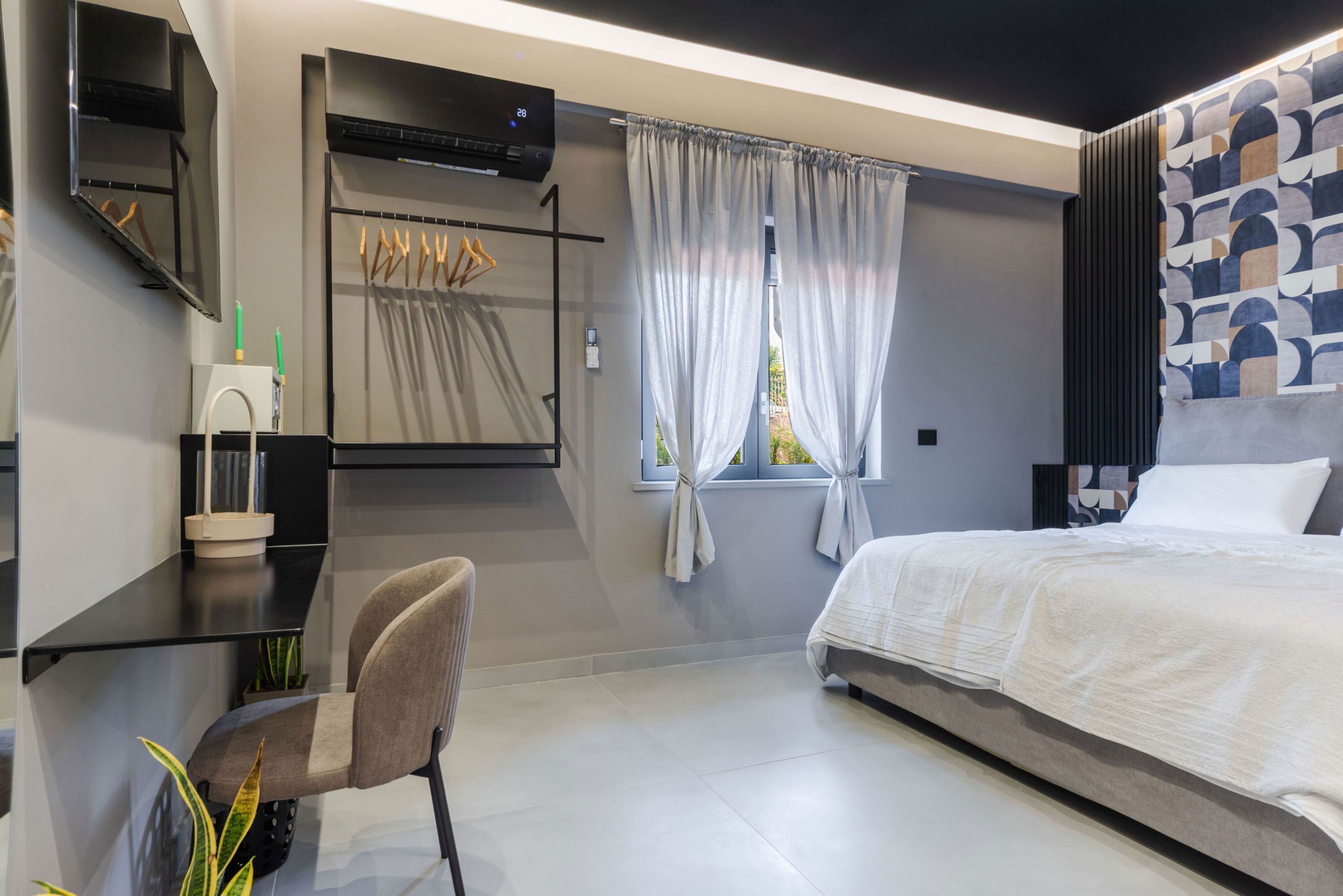 Un dormitorio moderno con cama, escritorio y silla. Se observa una ventana con cortinas, un aire acondicionado de pared y un diseño de pared estampado.