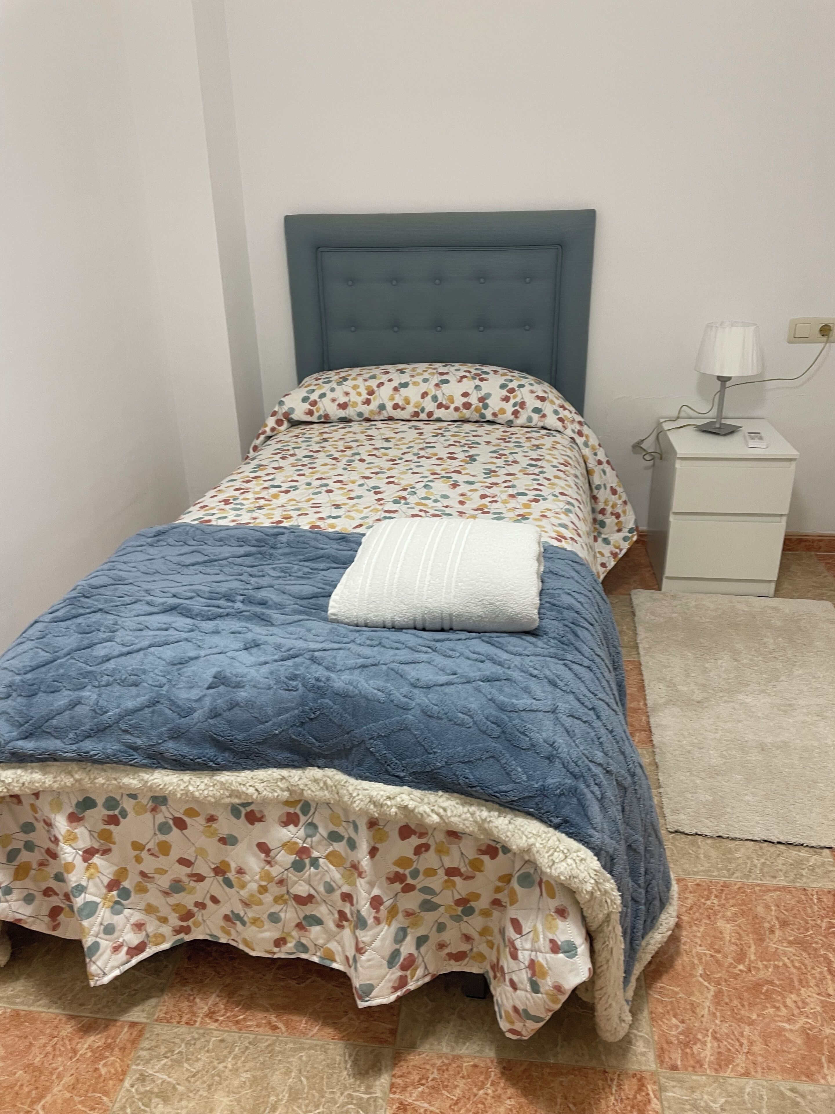 Un lit avec une couette bleue et une couvre-lit à motifs floraux, un oreiller blanc et une table de chevet avec une lampe.