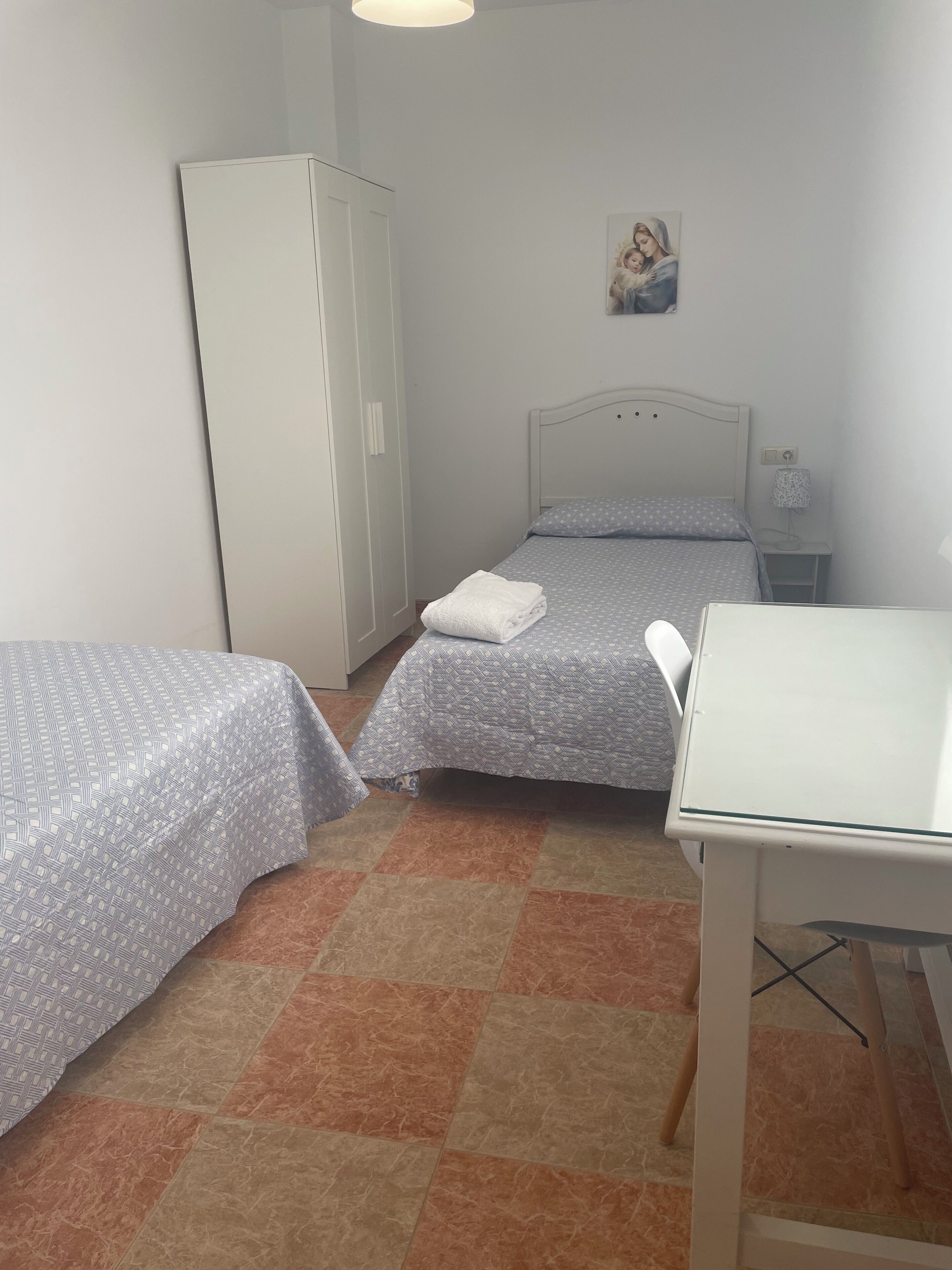 Una habitación pequeña y ordenada con dos camas individuales, un armario blanco, una mesita y una lámpara de pared.