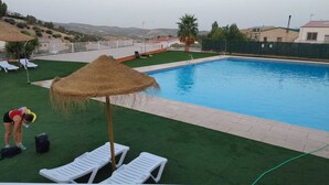 A swimming pool with a thatched umbrella, white lounge chairs, and a grassy area with a view of hills in the background. - El Mirador de Sierra Mágina II ( ideal for family travel) (Noalejo)