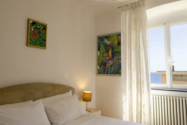 A hotel room with a bed, a nightstand with a lamp, two paintings on the wall, and a window with curtains.