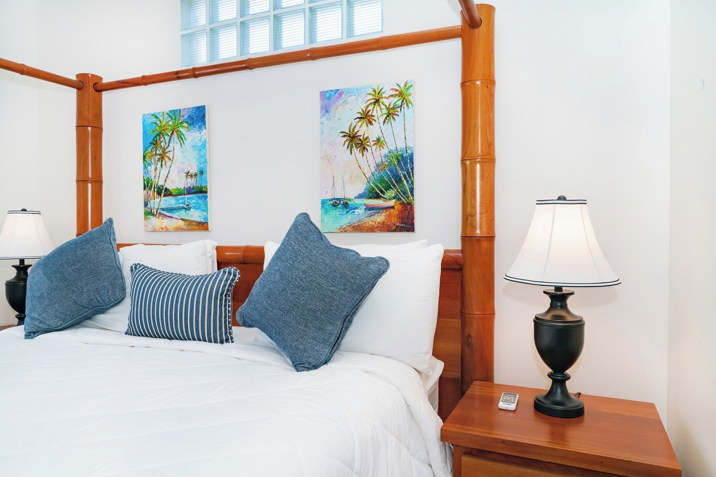 A bed with white bedding and blue pillows, a nightstand with a lamp, and two paintings of a beach scene on the wall.