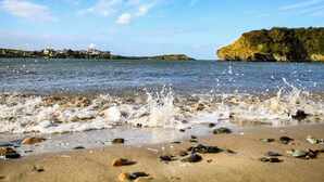 A beach with waves crashing onto the shore, scattered rocks, and a distant view of houses and cliffs. - 6 Gwelfor Estate (Cemaes Bay)