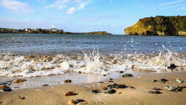 A beach with waves crashing onto the shore, scattered rocks, and a distant view of houses and cliffs. - 6 Gwelfor Estate (Cemaes Bay)