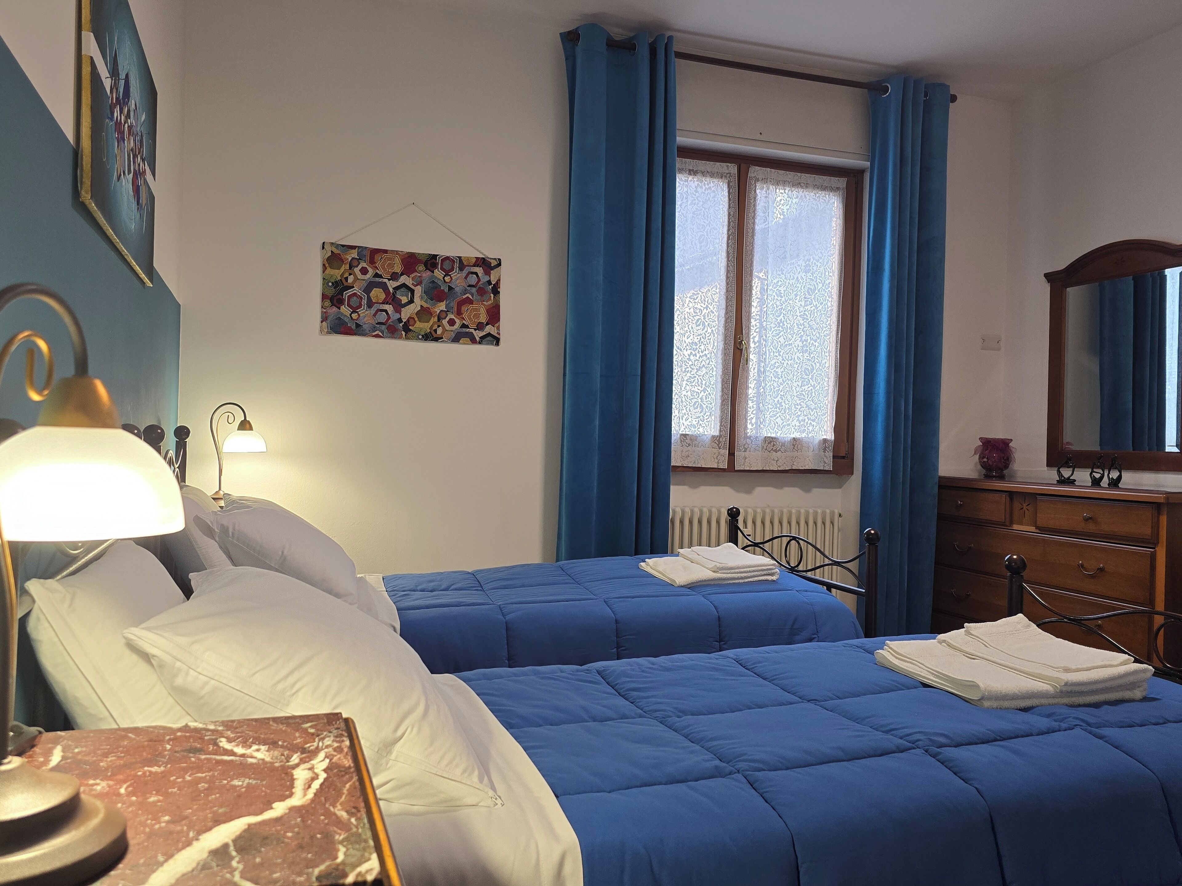 Ein Schlafzimmer mit zwei Betten, einem Nachttisch, einer Lampe, einer Kommode, einem Fenster mit Vorhängen und einem Wandbild.