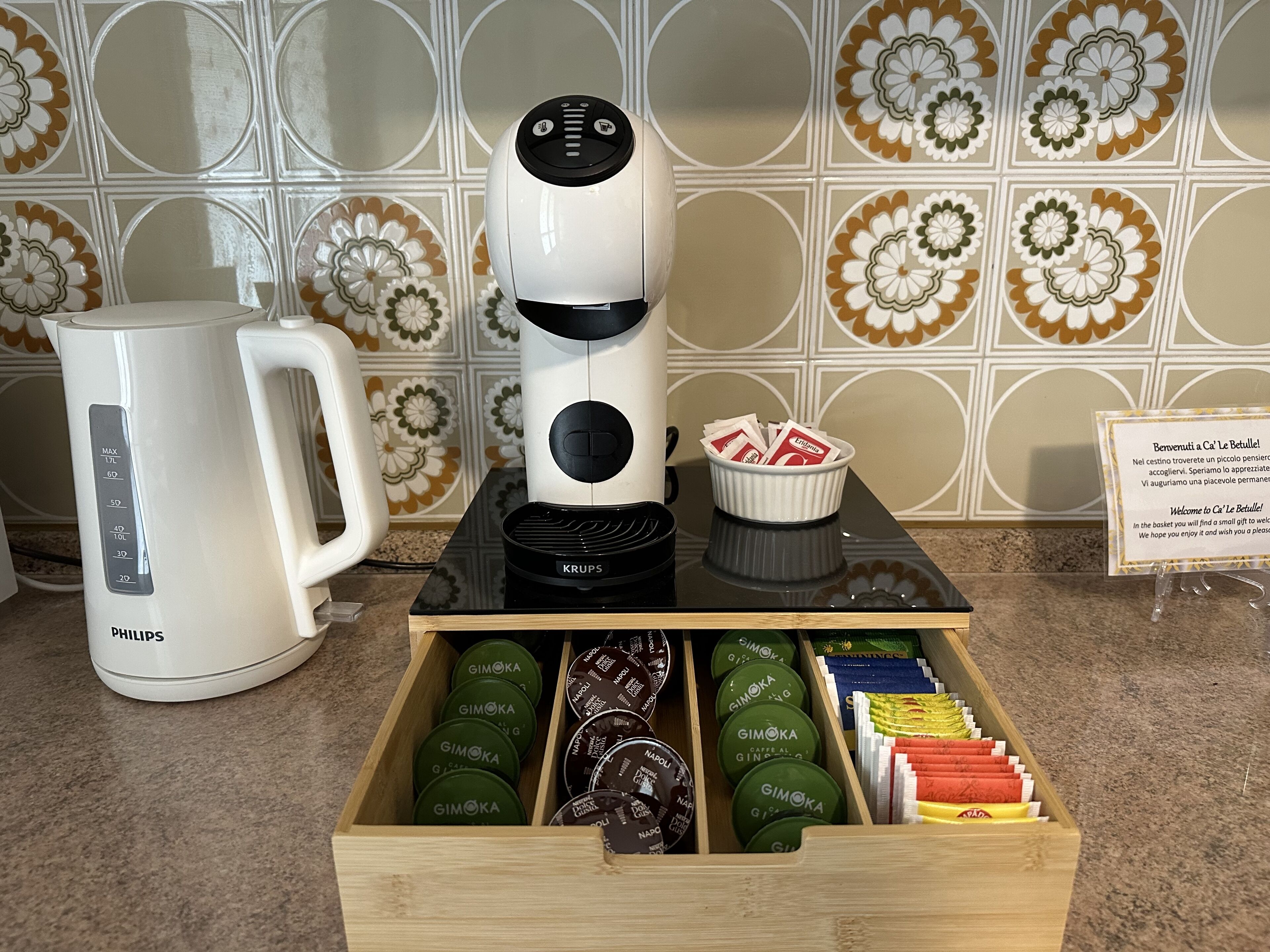A coffee maker, a kettle, and a basket with coffee pods on a table.