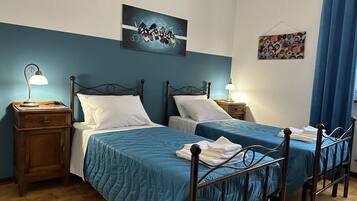 A double room with two beds, each with a wooden headboard and a blue bedspread. There are two bedside tables with lamps, and a painting is hung on the wall above the beds.