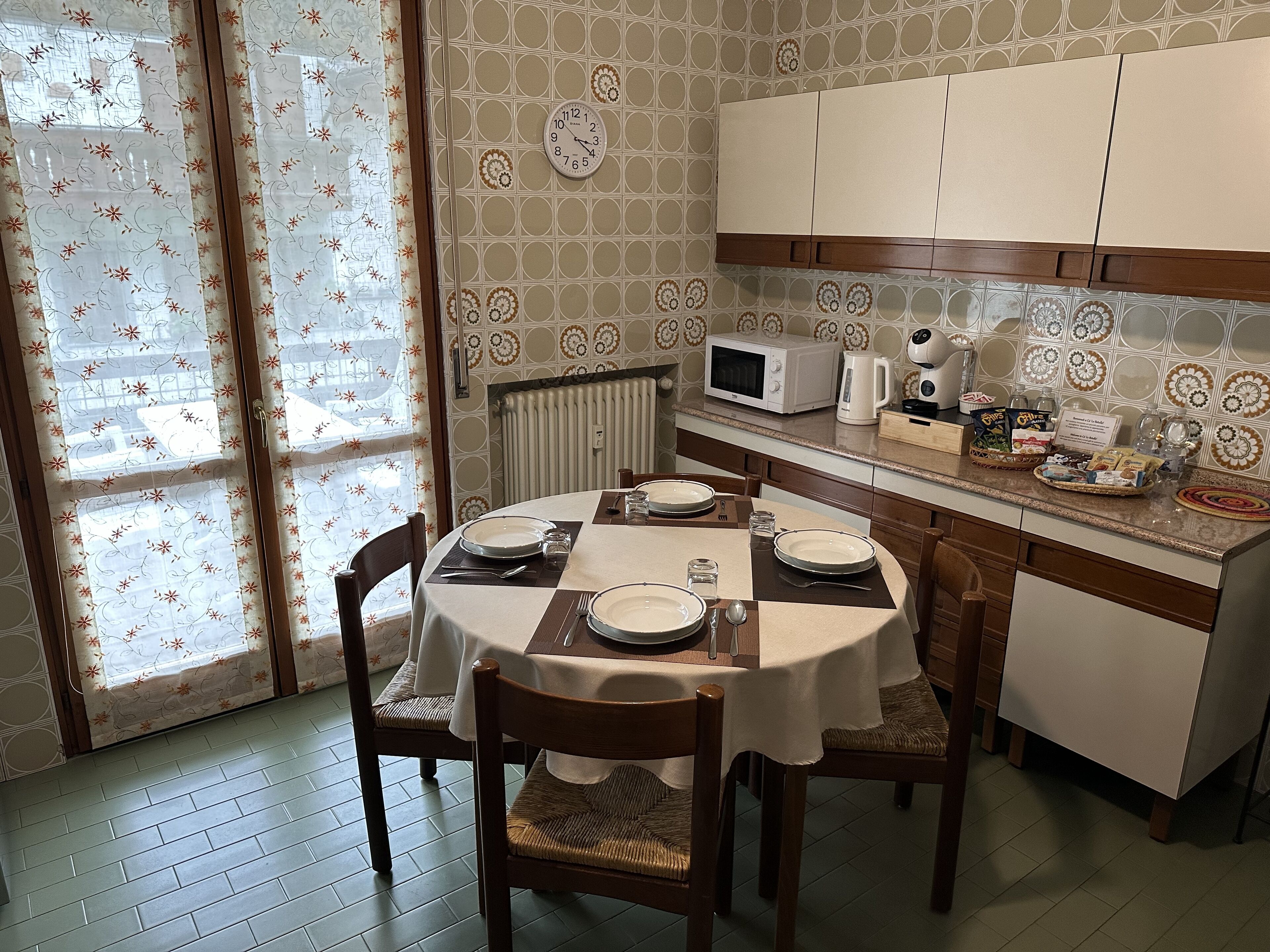 A dining area with a round table set for four, equipped with plates, cutlery, and glasses. There are wooden chairs with woven seats. A microwave and kettle are on the counter, and a small refrigerator is visible.