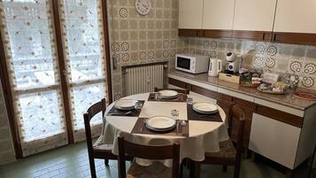 A dining area with a round table set for four, equipped with plates, cutlery, and glasses. There are wooden chairs with woven seats. A microwave and kettle are on the counter, and a small refrigerator is visible.