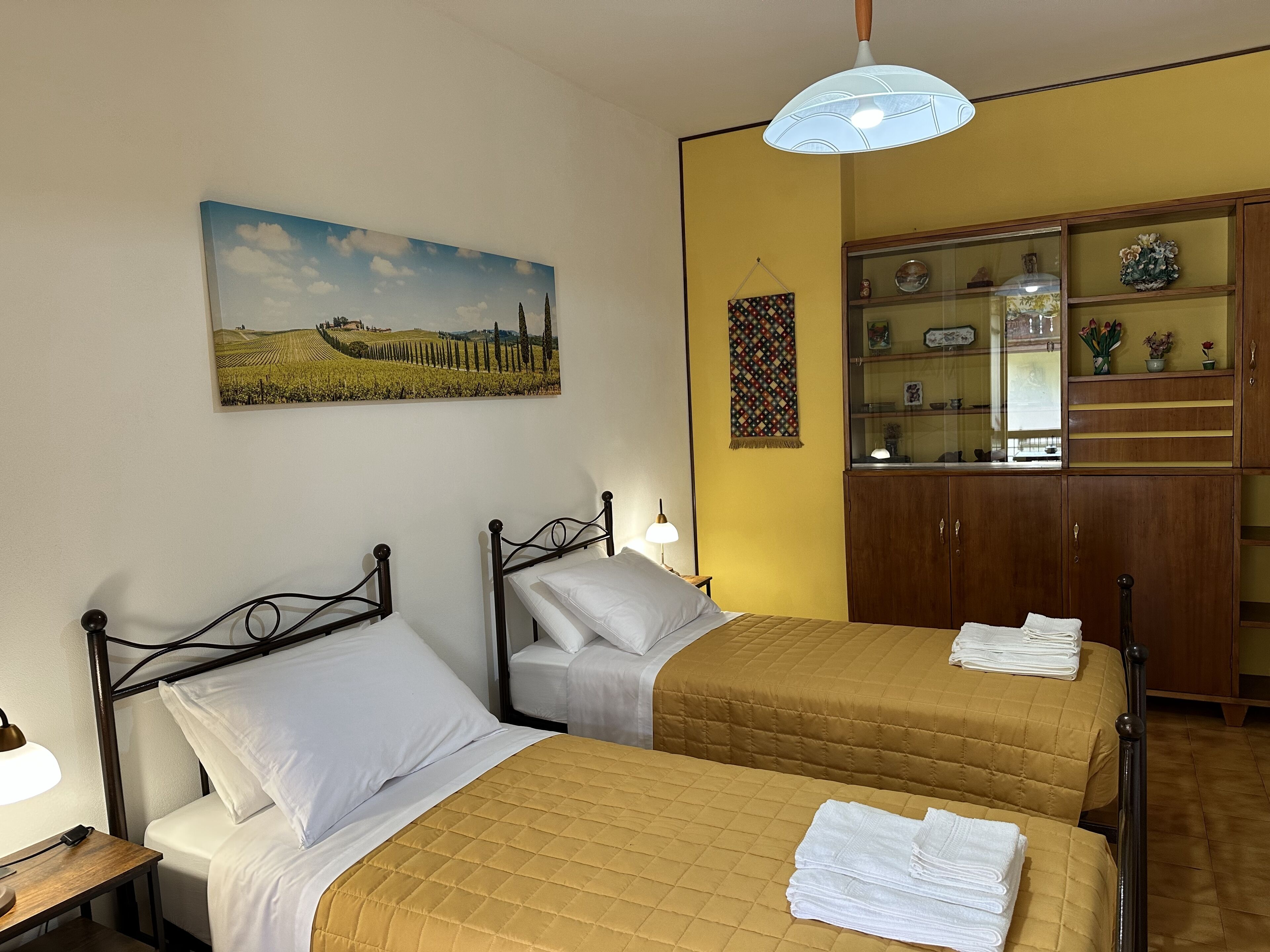A hotel room with two beds, a painting of a landscape, a wooden cabinet with glass doors, and a hanging tapestry.