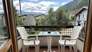Ein Balkon mit zwei weißen Plastikstühlen und einem Tisch, einer Satellitenschüssel und Blick auf Berge und Häuser.