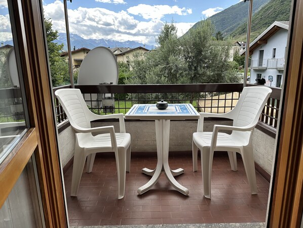 A balcony with two white plastic chairs and a table, a satellite dish, and a view of mountains and houses.
