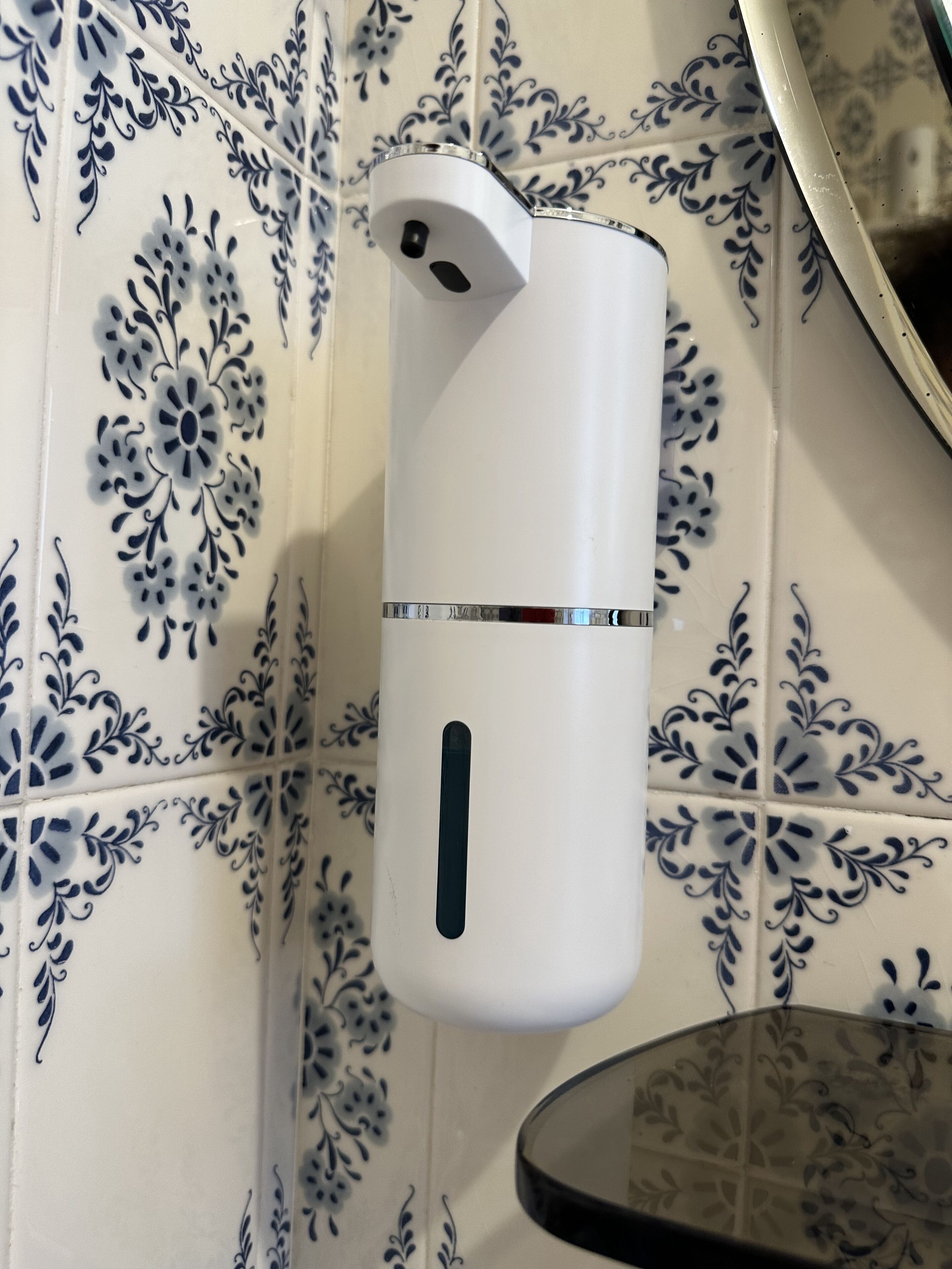 A white wall-mounted object with a metallic band, set against a patterned tile background.