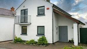 Uma casa moderna de dois andares com varanda, uma porta cinzenta e um pequeno jardim com plantas.