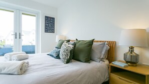 Cama bem arrumada com colcha listrada, almofadas verdes e estampadas, e mesa de cabeceira com abajur e um livro.