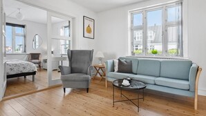 Een kamer met een lichtblauwe bank, een grijze fauteuil, een klein rond tafeltje met een theepot en een bed met een bloemenbedekking.